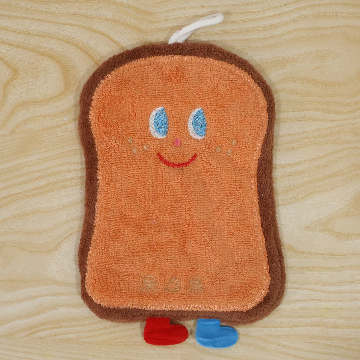 A brown cartoon bread slice towel with blue eyes, red smile, and two small feet, lying flat on a wooden surface.