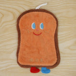 A brown cartoon bread slice towel with blue eyes, red smile, and two small feet, lying flat on a wooden surface.
