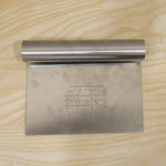 A stainless steel dough scraper with measurement markings and conversion chart sits on a light wooden surface.