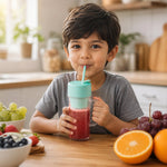 A young boy sips a red smoothie from a clear blender bottle with a mint green lid and straw in a kitchen setting.