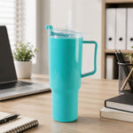 An aqua blue tumbler with a handle, clear lid, and straw sits on a wooden desk next to a laptop and office supplies.