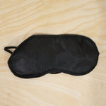 A black silk satin sleep mask with elastic straps lying flat on a light wooden surface.