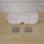 White plastic eyewear holder with two clear adhesive strips on a light wooden surface, against a white tiled wall.
