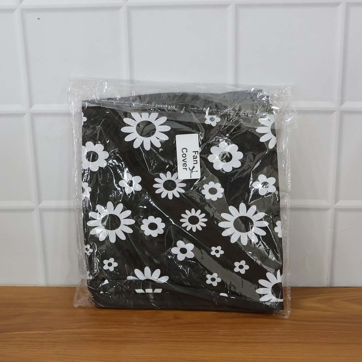 A folded black and white floral fan cover in clear plastic packaging, resting on a wooden surface.