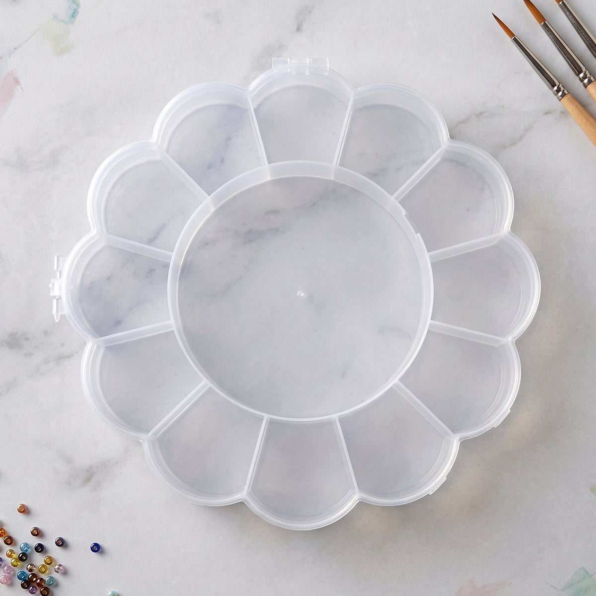 Empty clear flower-shaped plastic organizer box with 13 compartments on a white marble surface with paintbrushes nearby.