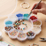 A hand using tweezers to pick a blue bead from a clear flower-shaped organizer box filled with various colorful beads and