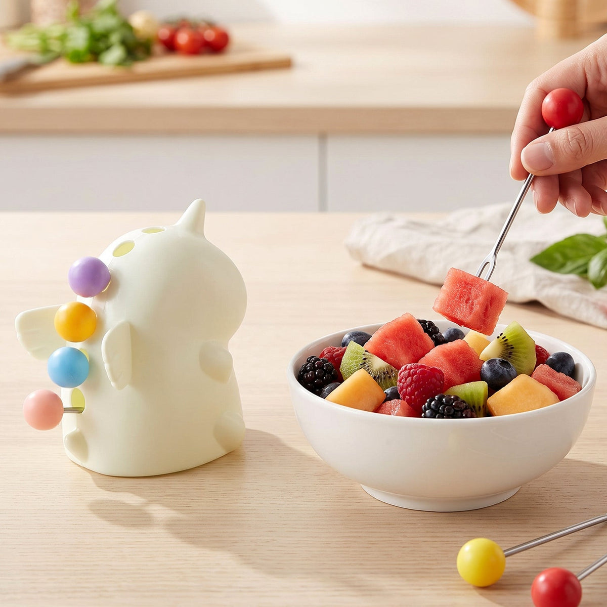 A hand uses a red-tipped fruit fork to pick up watermelon from a white bowl of mixed fruit next to a unicorn stand. – view 4
