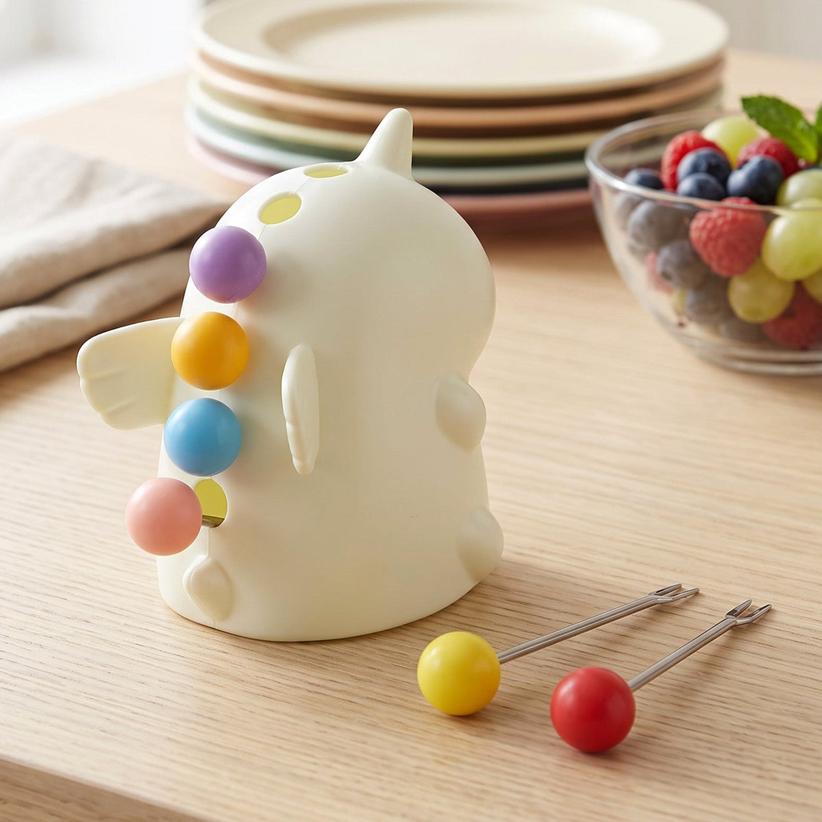 A cream unicorn stand with five colorful fruit forks on a wooden surface, with a bowl of fruit and stacked plates behind.