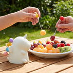 Two hands use colorful fruit forks to pick up melon and grapes from a white plate of mixed fruit outdoors.