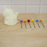 A cream unicorn stand sits beside six colorful fruit forks laid out on a light wooden table against a white tiled wall.