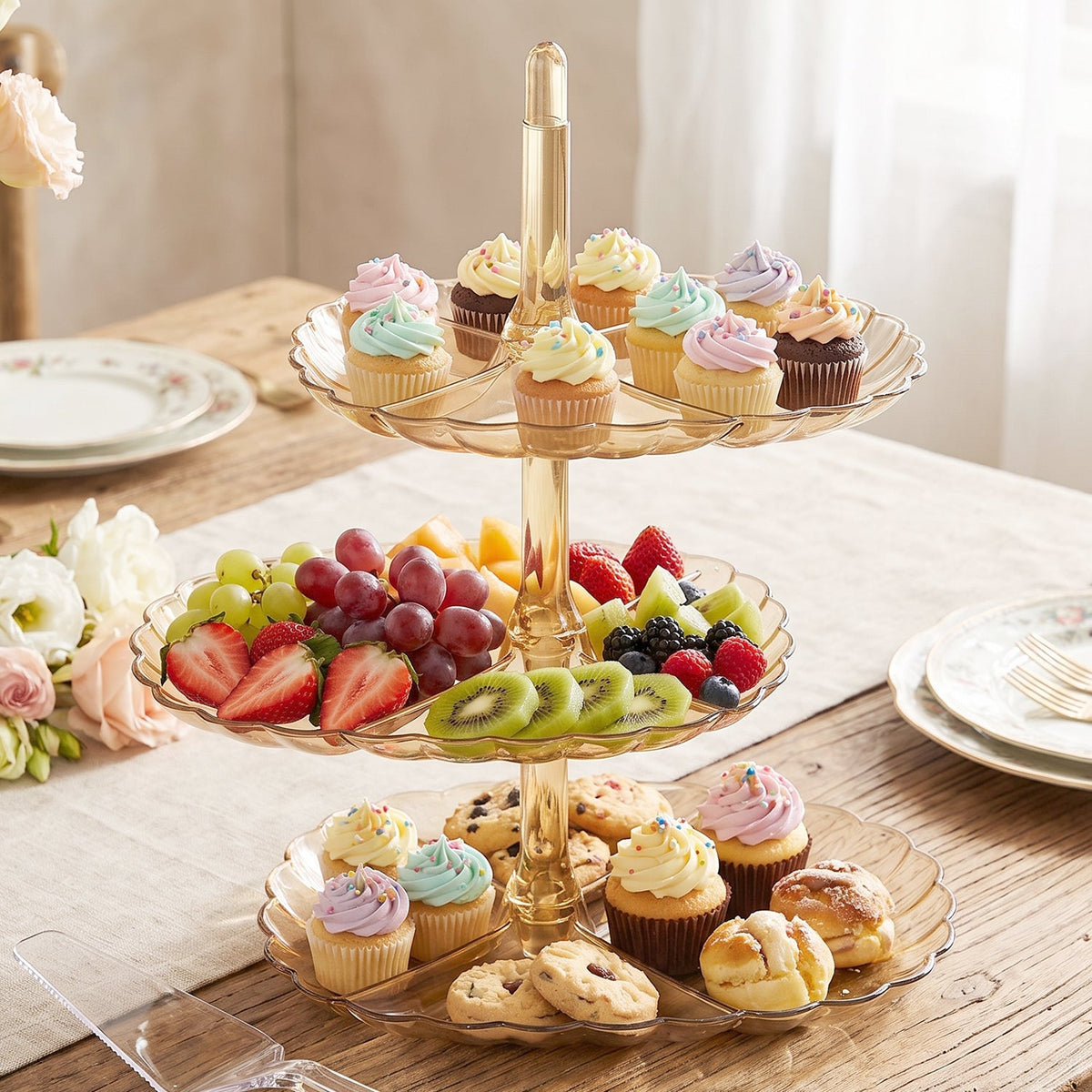 A three-tier amber acrylic stand displays an assortment of cupcakes, fresh fruit, and cookies on a wooden table.
