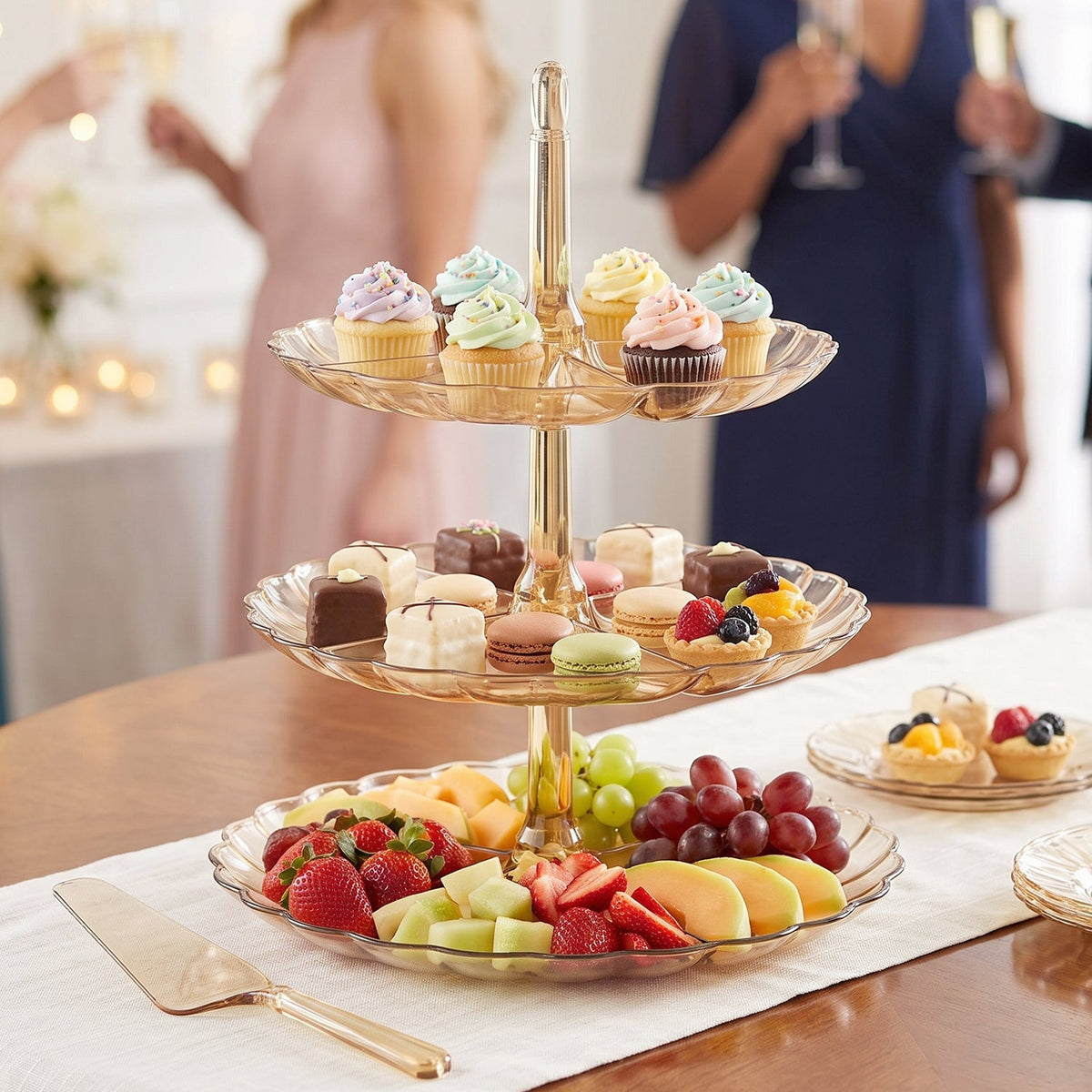 A three-level amber acrylic stand filled with cupcakes, pastries, and fruit is on a table with people in the background.