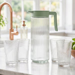 A woman pours water from a clear ribbed glass pitcher with a green lid into a matching glass tumbler on a light wooden