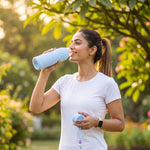 A smiling woman in a white t-shirt drinks from a light blue ribbed glass water bottle in a sunny outdoor setting.