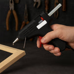 A hand holding a black and red glue gun, with a drop of hot glue dripping from the nozzle onto a wooden frame.