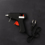 A black and red glue gun with a European plug and cord, neatly coiled, on a dark gray textured background.