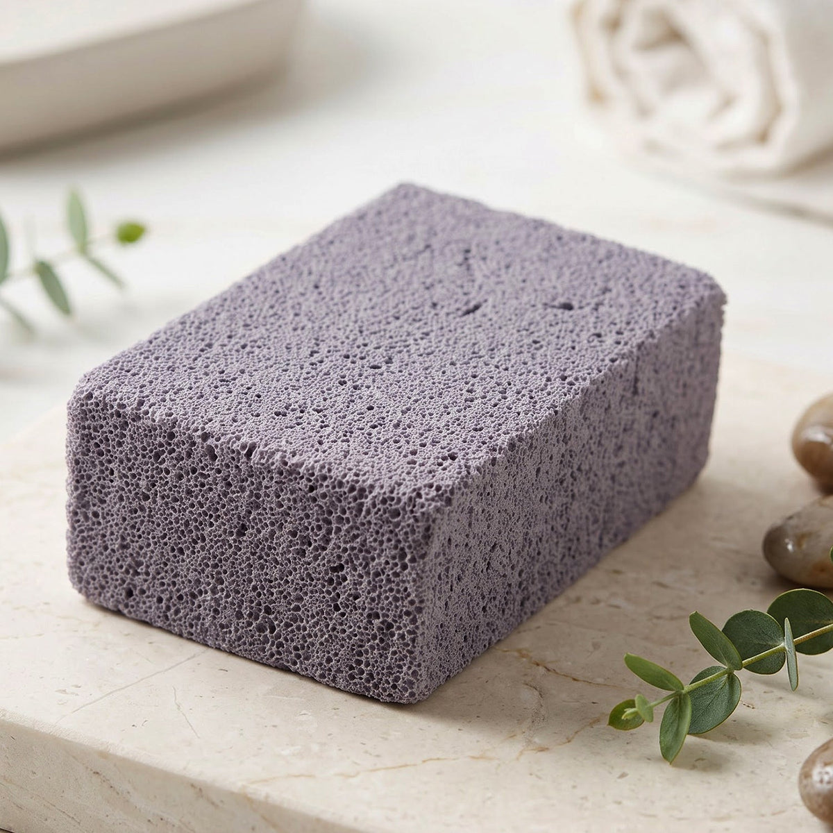 A single gray cleaning block with a porous texture resting on a light marble surface with green leaves and pebbles.