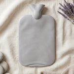A grey ribbed hot water bottle lies flat on a light fabric, with lavender sprigs and smooth white stones beside it.