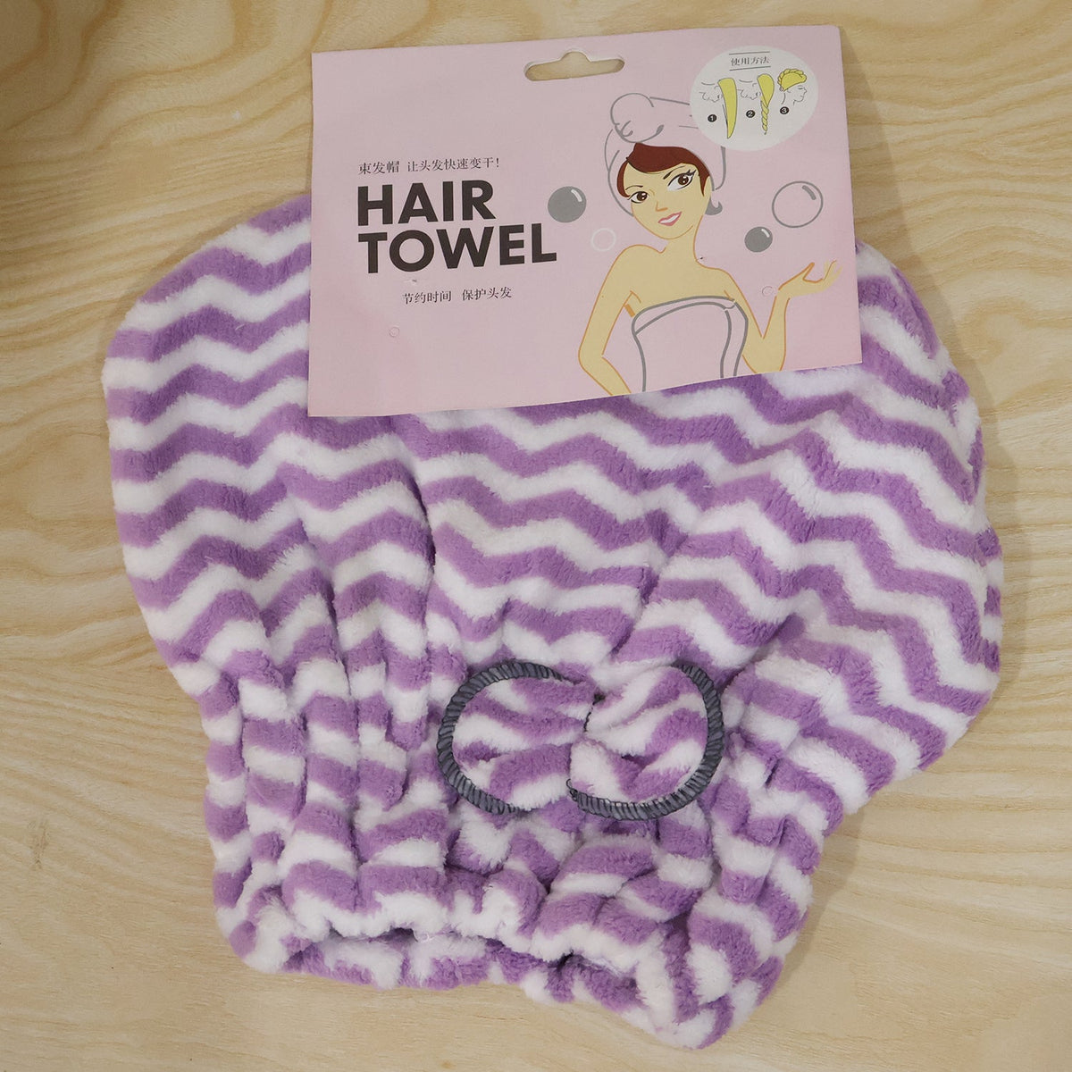 A purple and white chevron microfiber hair towel with a grey bow, displayed with its pink packaging on wood.