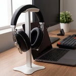 Black headphones resting on a white and silver stand, positioned on a wooden desk with a laptop and monitor.