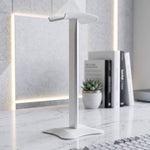 Silver headphone stand with a white base on a marble desk, with books and a plant in the soft-lit background.