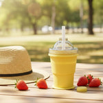 A yellow silicone cup with a straw and dome lid sits on a wooden table outdoors with strawberries and a straw hat.