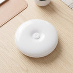 A white, round LED light with a central power button rests on a light wood desk next to a pink mat