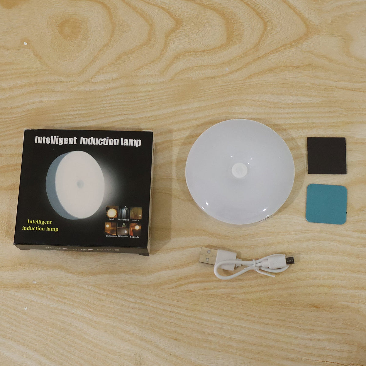 A black box labeled "Intelligent induction lamp," a white round LED light, a USB cable, and two adhesive pads on a wooden