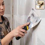 A person uses a stainless steel putty knife with a black and yellow handle to apply white putty to a wall.