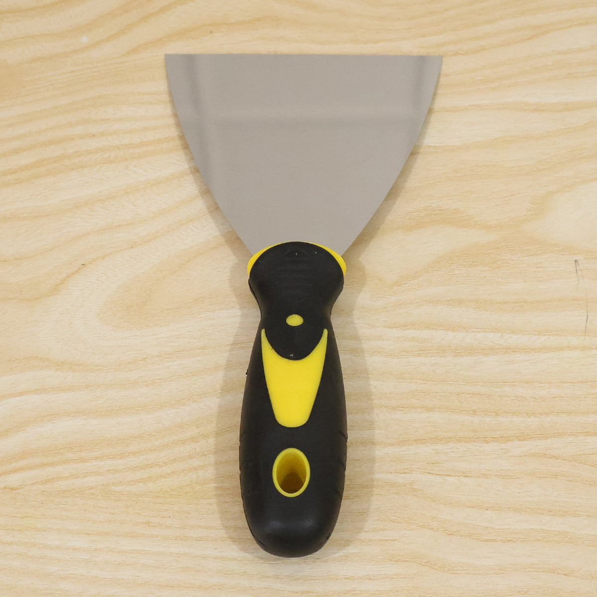 A stainless steel putty knife with a black and yellow handle is centered on a light wooden background.