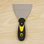 A stainless steel putty knife with a black and yellow handle is centered on a light wooden background.