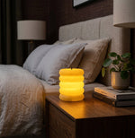 A single illuminated cream ribbed LED candle sits on a bedside table next to a bed in a dimly lit room.