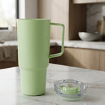 A light green travel tumbler with a handle stands on a marble counter next to its clear lid in a kitchen.