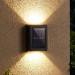 A black solar LED wall light illuminates a textured beige wall with warm light beams pointing up and down, with green