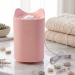 A pink cat-shaped mosquito lamp with a USB cable sits on a marble surface with decorative stones and folded blankets.