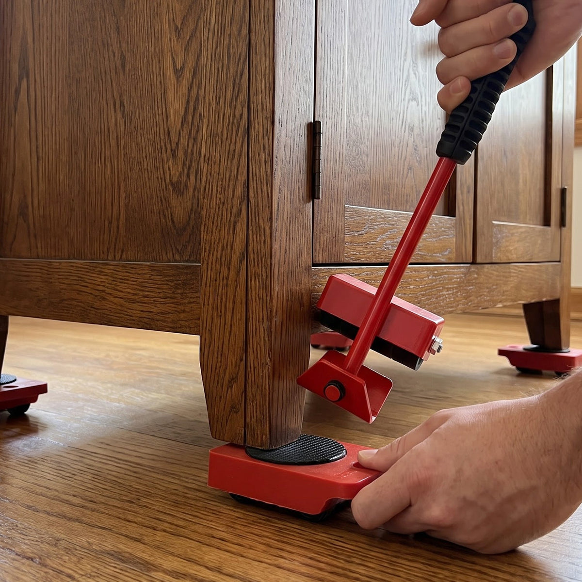 Hands operate the red furniture lifter to place a wheeled slider under a wooden cabinet leg on a hardwood floor.