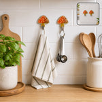 Two mushroom-shaped adhesive hooks hold a striped towel and measuring spoons on a white tiled kitchen wall.