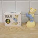 A yellow bear phone holder with blue overalls next to its product box on a wooden surface.