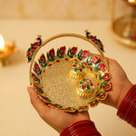 Hands holding a golden kumkum dish with a peacock handle and two lidded containers, with a blurred background.