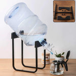 A large clear water bottle sits tilted on a black metal stand, dispensing water into a glass teapot.