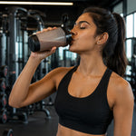 A woman in a black sports bra drinks from a grey and black shaker bottle in a gym.