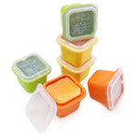 Six colorful square silicone molds with clear lids, some stacked, one open revealing frozen orange puree, on a white surface.