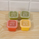 Four square silicone molds in green, red, and yellow with clear lids, arranged on a light wooden surface against a white