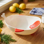 A white whale-shaped rice drainer bowl with red accents sits on a wooden counter next to lemons and fresh dill.