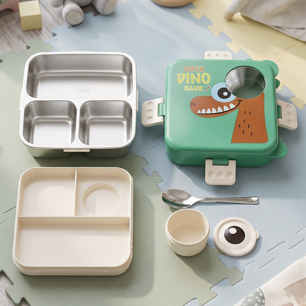 The green dinosaur lunchbox components are neatly arranged on a soft play mat, including the outer box, inner tray, and