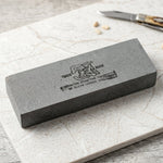 Gray rectangular sharpening stone with black text and tiger logo on a white marble surface next to a small knife.