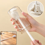 A hand holds a clear glass bottle being cleaned by a white silicone brush, with a close-up of the brush head.