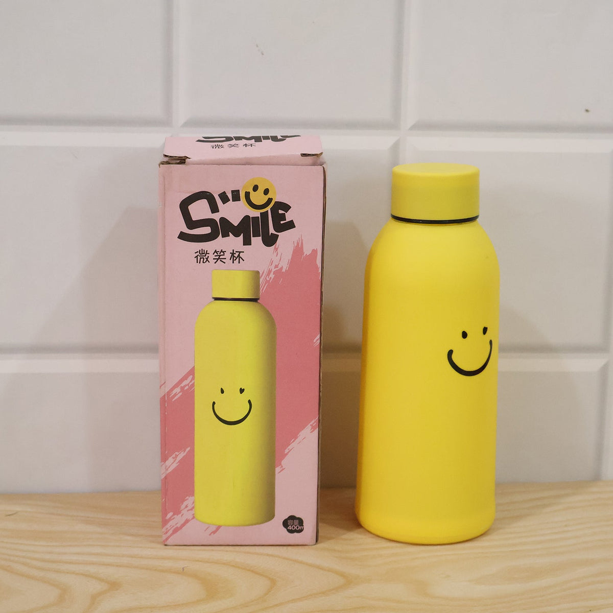 A yellow glass water bottle with a smiley face stands next to its pink and white packaging box.