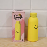 Yellow glass water bottle with a smiley face next to its pink packaging box, both featuring the same design.