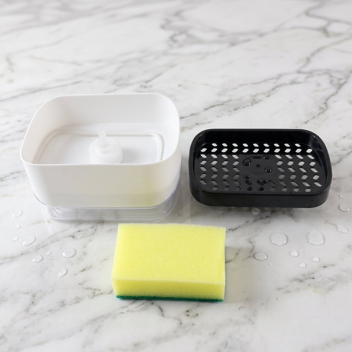 The white soap dispenser base, black perforated top, and yellow sponge are separated on a white marble surface with water